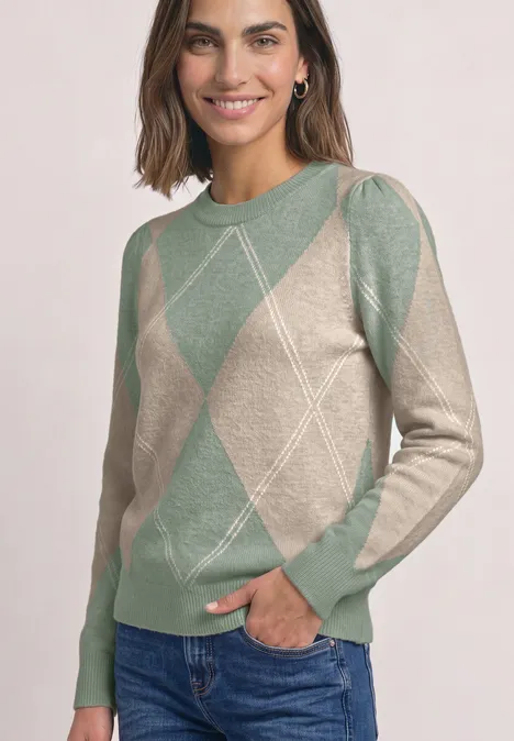 Lächelnde Frau in beige-grünem Rautenmuster-Pullover mit langen Ärmeln und Jeans, helles Studio.