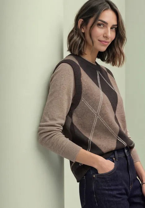 Bezrękawnik z okrągłym dekoltem i wzorem argyle sanded amber mel. Kobieta opiera się o ścianę, ma na sobie brązowy sweter w romby i ciemne jeansy, swobodna poza.