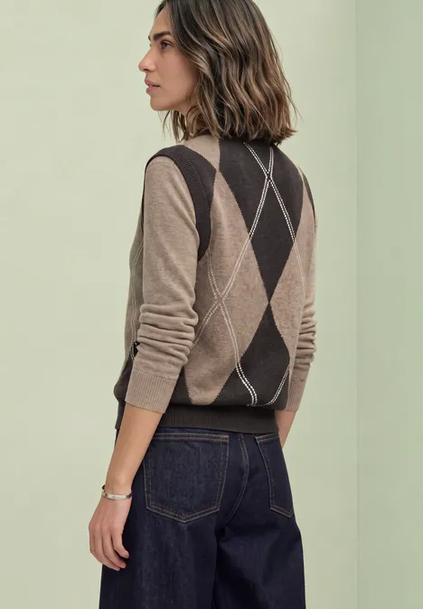 Bezrękawnik z okrągłym dekoltem i wzorem argyle sanded amber mel. Profil kobiety w argyle’owym sweterku bez rękawów i ciemnych jeansach na jasnym zielonym tle.
