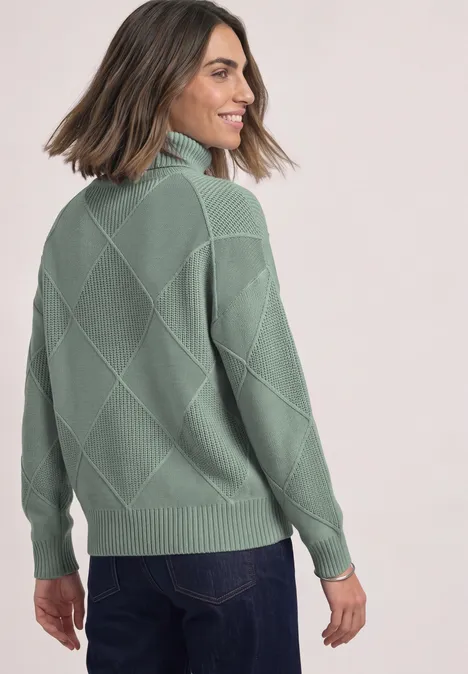 Vrouw kijkt over haar schouder, draagt groene trui met ruitmotief en een spijkerbroek.