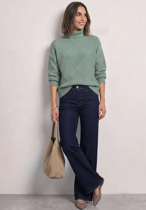 Vrouw in groene trui, donkere wijde jeans en beige tas voor een neutrale achtergrond.