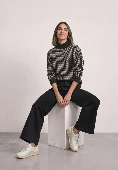 Pullover mit Turtle Neck und Streifenmuster black coffee Junge Frau in gestreiftem Pullover und schwarzen Hosen sitzt entspannt auf einem weißen Würfel vor hellem Hintergrund.