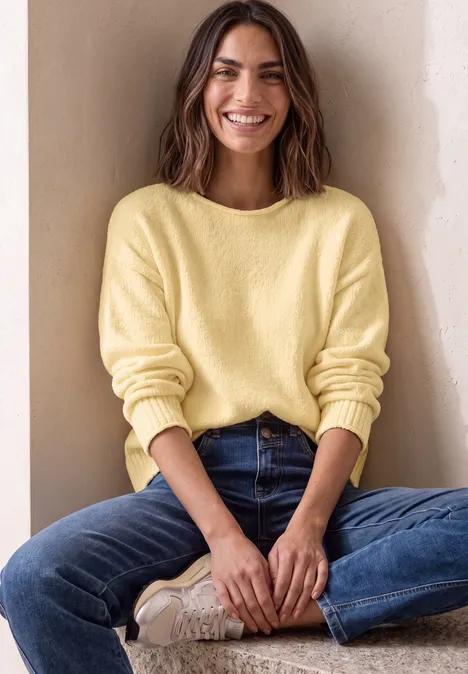 Chenille-Pullover mit U-Boot-Ausschnitt sunbeam yellow Eine Frau sitzt lächelnd im Schneidersitz, trägt gelben Pullover und blaue Jeans, vor beiger Wand.