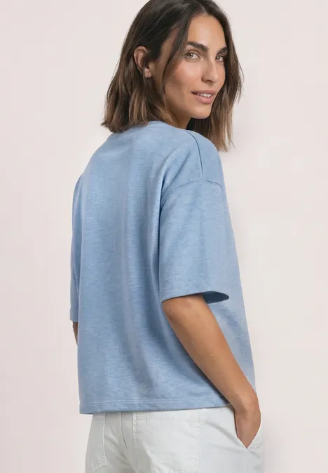 Frau blickt über die Schulter, trägt hellblaues T-Shirt und weiße Hose, eine Hand in der Tasche.