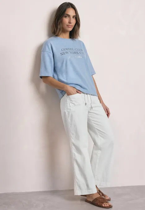 Eine Frau lehnt an einer hellen Wand, trägt weiße, weite Hose, blaues T-Shirt und braune Sandalen.