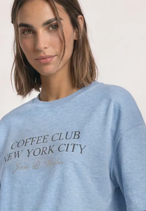 Frau trägt hellblaues Sweatshirt mit Aufschrift "Coffee Club New York City", lächelnd
