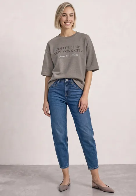 Lächelnde Frau mit kurzem blondem Haar, grauem T-Shirt, blauen Jeans und grauen Ballerinas.
