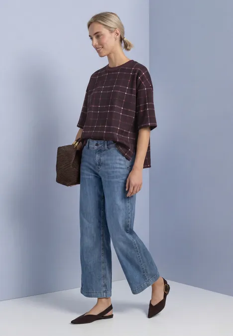Frau steht vor grauem Hintergrund, trägt karierte Bluse, weite Jeans, schwarze Ballerinas, braune Clutch.