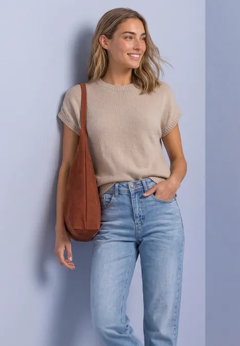 Mouwloze trui met ronde hals light sepia sand Eine freundlich lächelnde Frau in beigem Kurzarmshirt, hellblauen Jeans und brauner Tasche vor hellblauem Hintergrund.