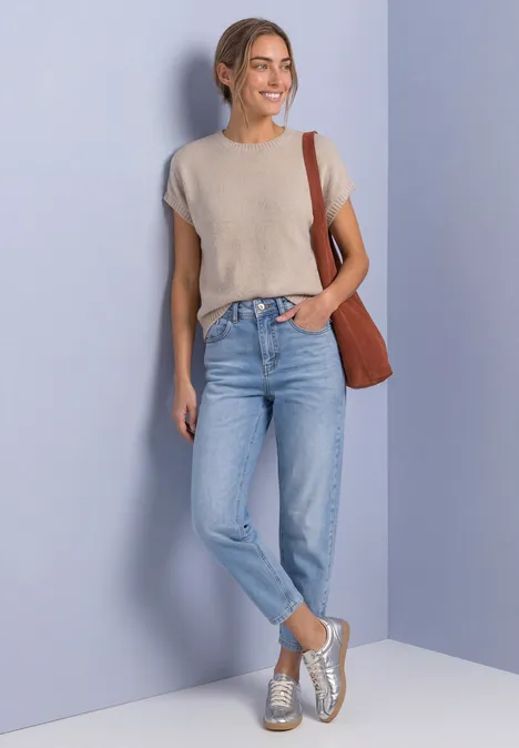 Mouwloze trui met ronde hals light sepia sand Eine Frau lächelt vor violettem Hintergrund, trägt graues T-Shirt, Jeans, silberne Sneaker und eine braune Tasche.