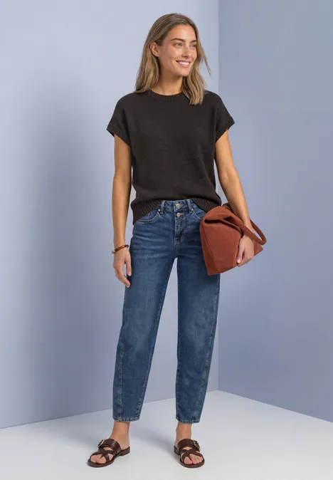 Mouwloze trui met ronde hals black coffee Eine Frau trägt schwarzes T-Shirt, blaue Jeans, braune Sandalen und hält eine rostfarbene Tasche vor hellem Hintergrund.