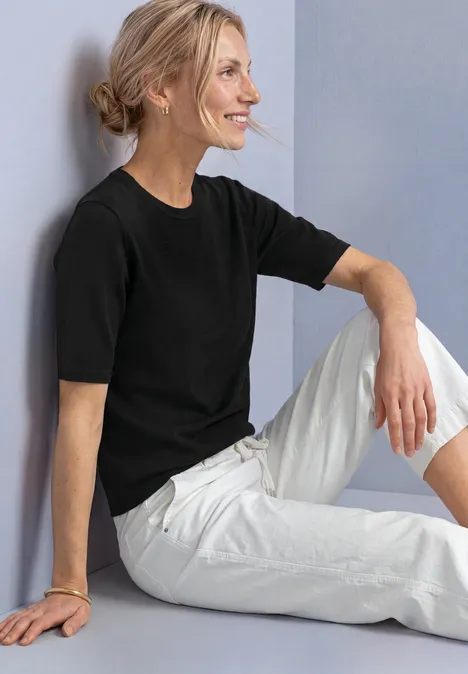 T-shirt met korte mouwen en ronde hals in effen kleur Black Een glimlachende vrouw in een zwart shirt en witte broek zit ontspannen.