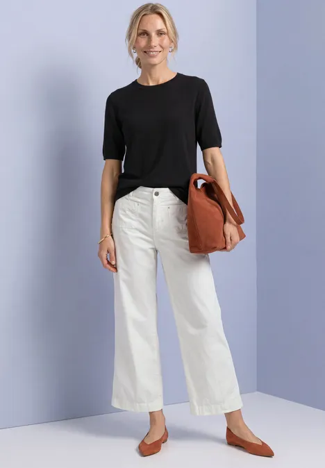 T-shirt met korte mouwen en ronde hals in effen kleur Black Vrouw in zwart T-shirt, witte wijde jeans en bruine ballerina’s met grote bruine tas voor paarse muur.