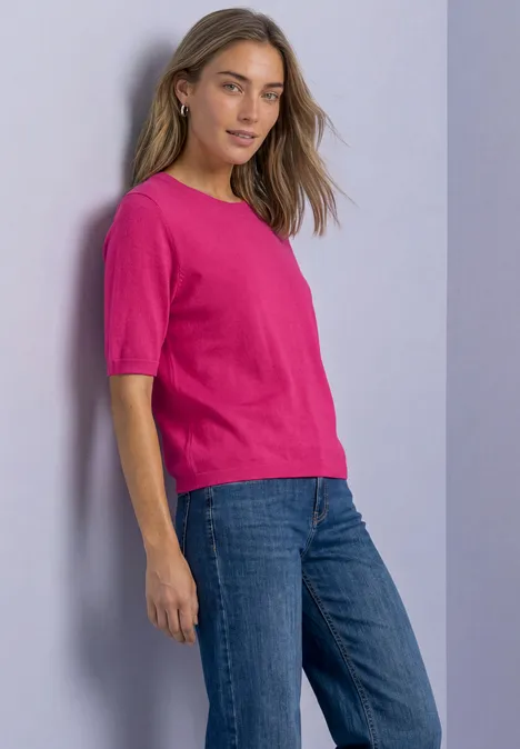 Een vrouw staat zijwaarts in een roze T-shirt met korte mouwen en een blauwe spijkerbroek.