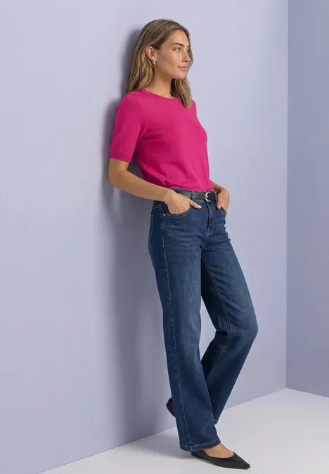 Een vrouw leunt tegen de muur met een roze shirt, blauwe jeans en zwarte ballerina’s.