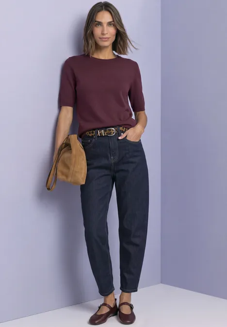 Vrouw met paarse kortemouwtrui, donkere jeans, bruine ballerina’s en tas voor een lila muur.