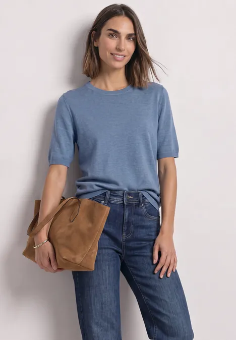 Een vrouw draagt een blauw shirt met korte mouwen, een spijkerbroek en houdt een bruine tas vast.