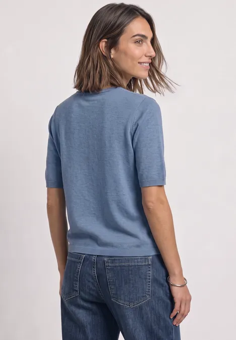 Vrouw van achteren in blauw t-shirt en spijkerbroek, korte mouwen, glimlachend, kijkt opzij.