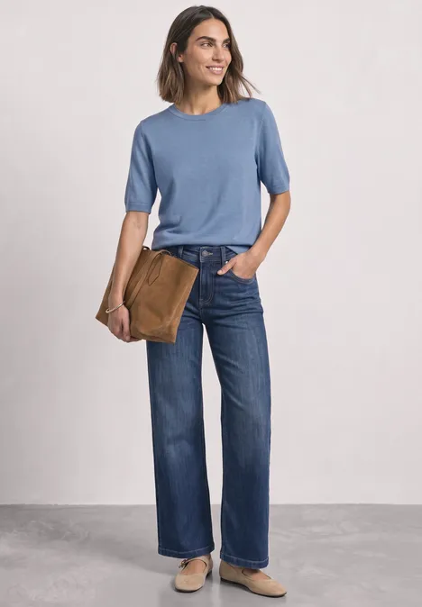 Een vrouw draagt een blauw T-shirt, wijde jeans en houdt nonchalant een bruine tas onder haar arm.