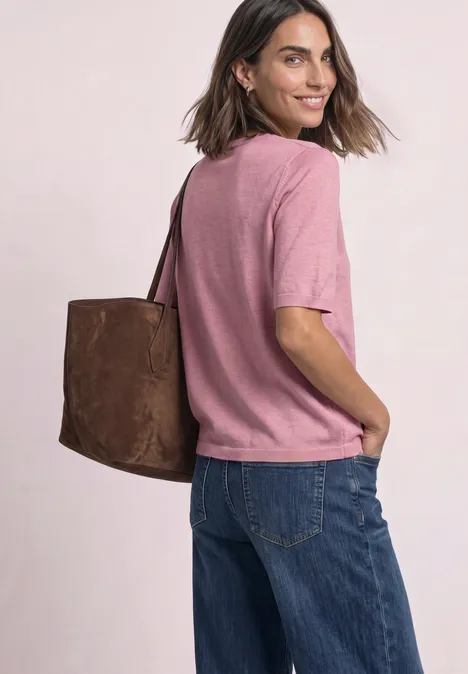 Vrouw in roze shirt en blauwe spijkerbroek met bruine tas kijkt over haar schouder.
