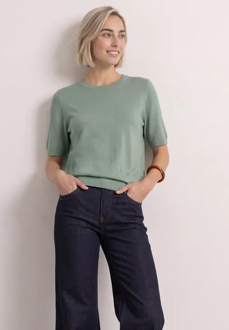 T-shirt met korte mouwen en ronde hals in effen kleur eucalyptus green Vrouw met kort blond haar, groene top met korte mouwen en wijde donkere jeans voor een lichte muur.