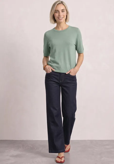 T-shirt met korte mouwen en ronde hals in effen kleur eucalyptus green Vrouw in groen T-shirt en wijde donkere jeans, ontspannen staand met handen in zakken voor een lichte achtergrond.