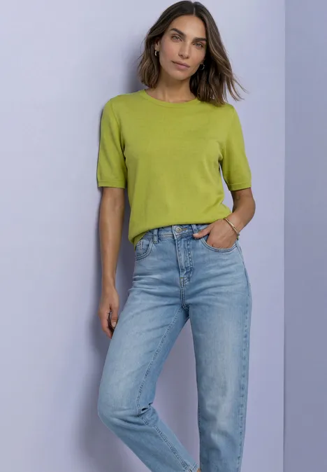 T-shirt met korte mouwen en ronde hals in effen kleur apple lime Vrouw voor paarse achtergrond in lichtgroen shirt en blauwe jeans, hand in zak, ontspannen houding.