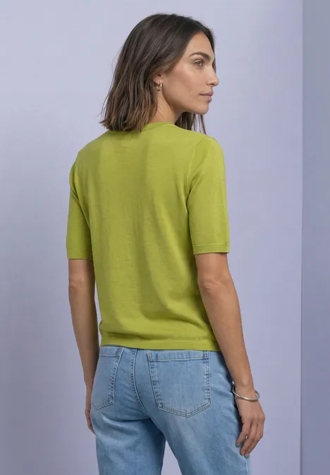 T-shirt met korte mouwen en ronde hals in effen kleur apple lime Een vrouw staat met haar rug naar de camera in een groen shirt met korte mouwen en een blauwe jeans voor een lichte muur.