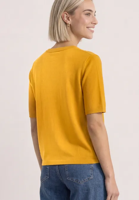 T-shirt met korte mouwen en ronde hals in effen kleur saffron yellow Vrouw van achteren in mosterdkleurige korte mouw top en blauwe jeans, glimlachend opzij kijkend.