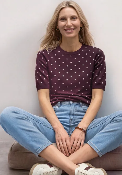Trui met korte mouwen, ronde hals en patroon rustic rouge Blonde vrouw zit in kleermakerszit voor een witte muur, draagt een paarse top met stippen en blauwe jeans.