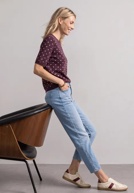 Trui met korte mouwen, ronde hals en patroon rustic rouge Vrouw leunt zijwaarts tegen een stoel, draagt paars gestipt shirt, lichte jeans en witte loafers.