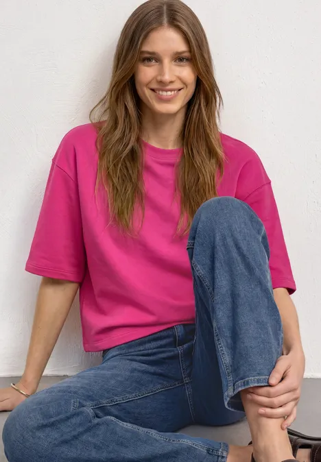 Sweatshirt met korte mouwen en ronde hals magenta dream Eine lächelnde Frau in pinkem T-Shirt und Jeans sitzt vor weißem Hintergrund.