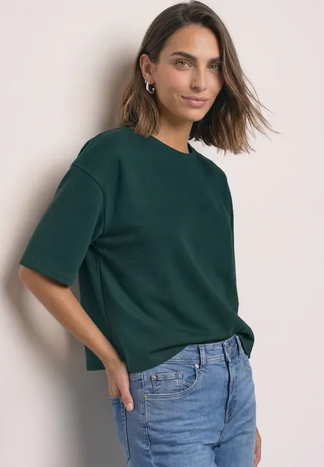 Sweatshirt met korte mouwen en ronde hals shadow teal Eine Frau mit braunem Haar trägt ein dunkelgrünes T-Shirt und blaue Jeans vor hellem Hintergrund.