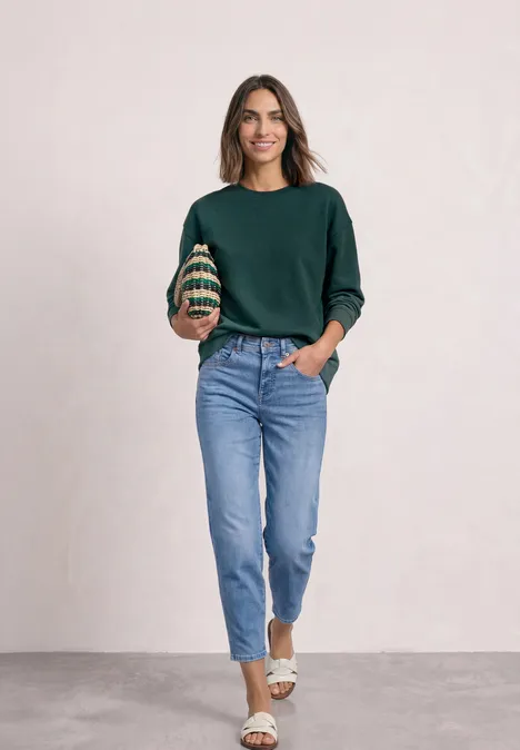 Sweatshirt met korte mouwen en ronde hals shadow teal Frau in grünem Pullover, blauen Jeans und weißen Sandalen steht vor neutralem Hintergrund und lächelt.