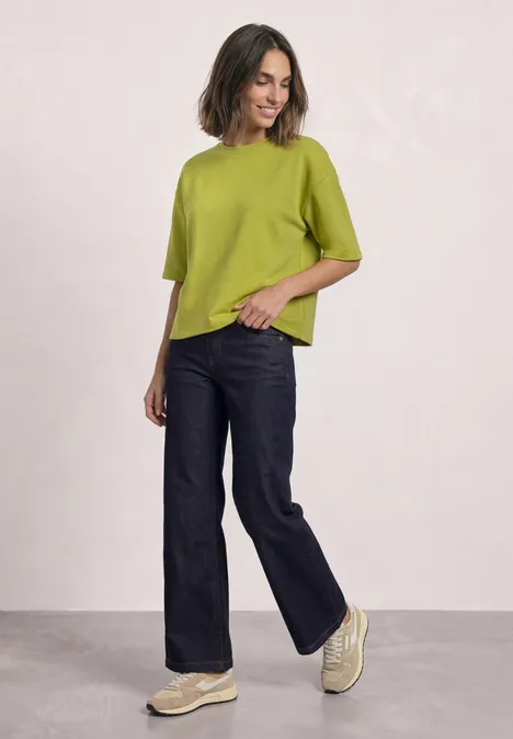 Sweatshirt met korte mouwen en ronde hals apple lime Eine Frau in einer grünen Bluse, weiten Jeans und Plateau-Sneakern posiert vor hellem Hintergrund.