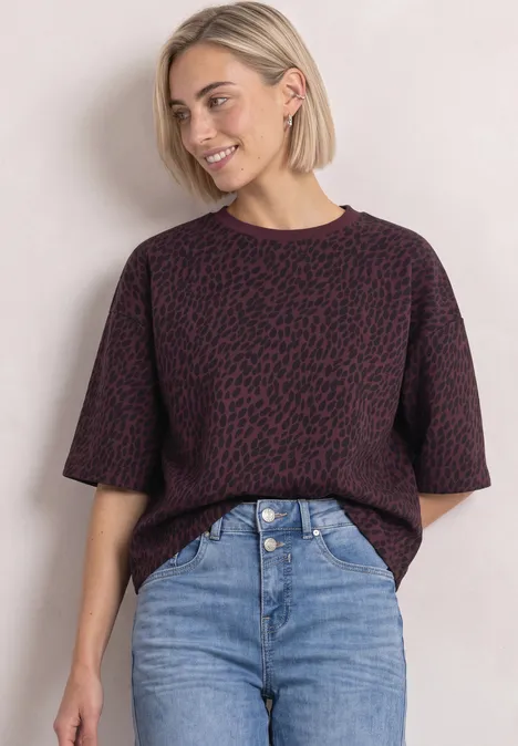Bluza z krótkim rękawem i okrągłym dekoltem rustic rouge Młoda kobieta z blond bobem ma na sobie krótki fioletowy wzorzysty sweter i jeansy z wysokim stanem na jasnym tle.