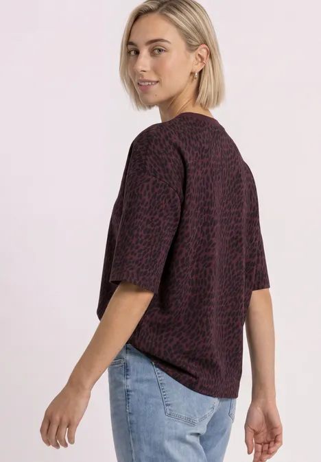 Bluza z krótkim rękawem i okrągłym dekoltem rustic rouge Kobieta w fioletowo-czerwonej koszulce w wzory i dżinsach spogląda przez ramię.