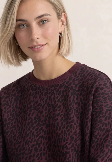 Bluza z krótkim rękawem i okrągłym dekoltem rustic rouge Kobieta o blond włosach ma bordowy sweter z czarnym motywem zwierzęcym.