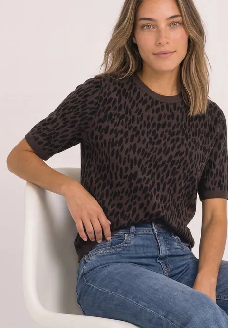 Jacquardtrui met ronde hals en luipaardprint molten earth Vrouw zit op witte stoel, draagt donkerbruin leopard-print truitje en blauwe jeans.