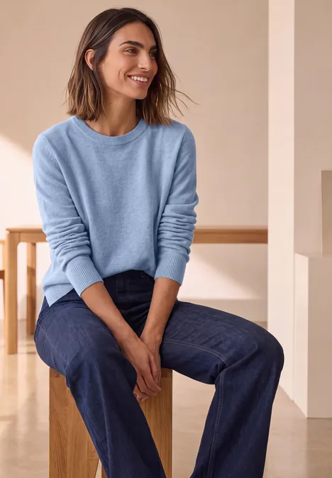 Pull à manches longues et col bateau northern blue mel. Femme en pull bleu et jean, assise sur un tabouret dans une pièce lumineuse, souriante.