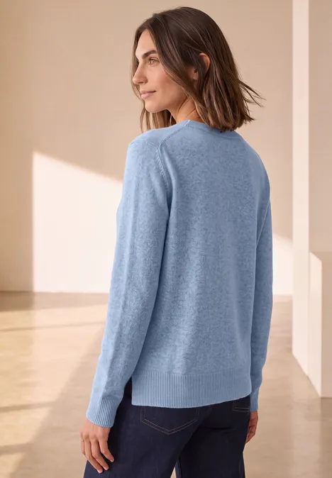 Pull à manches longues et col bateau northern blue mel. Femme dans une pièce claire, pull bleu clair et jean, regard par-dessus l’épaule, profil latéral.