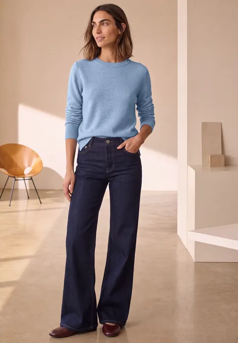 Pull à manches longues et col bateau northern blue mel. Femme dans un intérieur épuré portant un pull bleu clair, un jean évasé foncé et des chaussures marron.