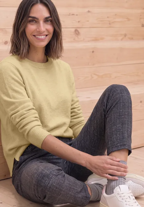 Pull à manches longues et col bateau sunbeam yellow mel. Une femme souriante, assise devant un mur en bois, porte un pull jaune, un pantalon gris et des baskets blanches.