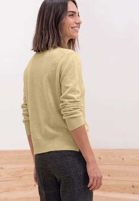 Pull à manches longues et col bateau sunbeam yellow mel. Femme de dos portant un pull clair et un pantalon foncé, devant un mur clair.