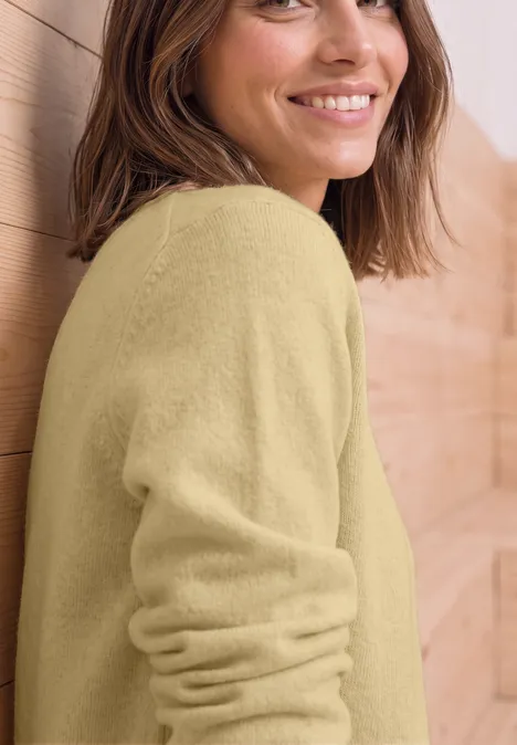 Pull à manches longues et col bateau sunbeam yellow mel. Une femme souriante en pull clair appuyée de manière détendue contre un mur en bois.