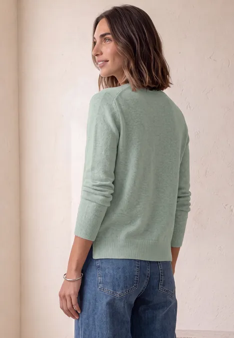Pull à manches longues et encolure bateau eucalyptus green mel. Une femme de profil porte un pull vert clair et un jean bleu, regardant par-dessus son épaule sur un fond neutre.
