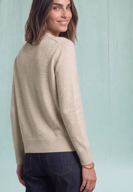 Pull à manches longues et col bateau clay sand mel. Femme en pull beige et jean regarde par-dessus son épaule, le visage rayonnant d’un léger sourire.