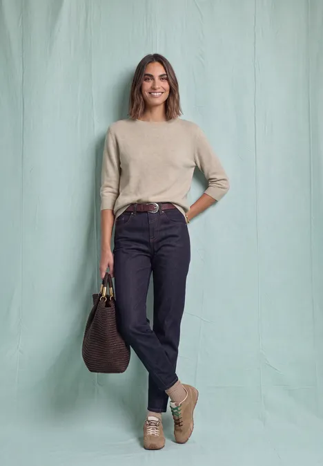 Pull à manches longues et col bateau clay sand mel. Femme en pull beige, jean noir, bottines claires et grand sac marron devant un fond pastel