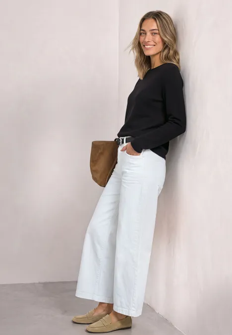 Pullover mit Boat Neck und Rollkanten Black Eine junge Frau lehnt lächelnd an einer hellen Wand, trägt schwarzen Pullover und weiße Jeans, hält eine braune Tasche.