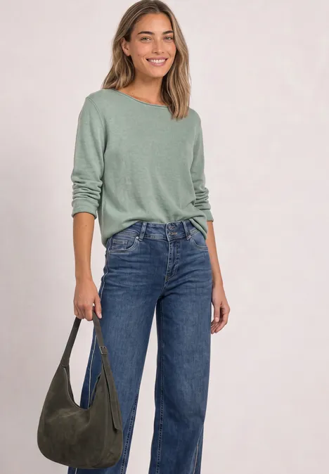 Une femme portant un t-shirt vert-gris à manches longues, un jean bleu et un sac vert, souriante.